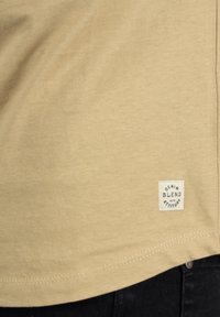 Svijetlosmeđi majica s mekanom teksturom, zaobljenim izrezom i malom izvezenom oznakom s natpisom "Denim Blend with Attitude" uvedenom na rubu.