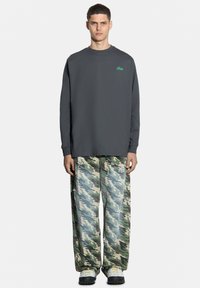 Szara koszula z długim rękawem z zielonym logo, w połączeniu z luźnymi spodniami w kamuflażowy wzór w odcieniach zieleni i beżu oraz szarymi trampkami.