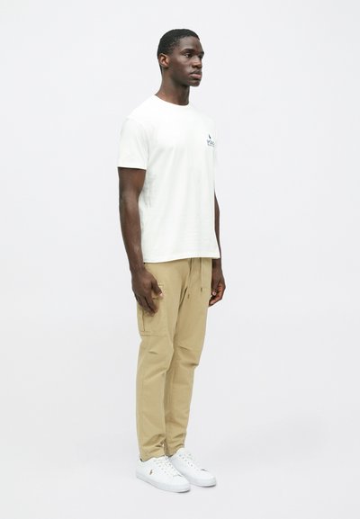 Λευκό κοντομάνικο μπλουζάκι με μπλε λογότυπο, μπεζ cargo παντελόνι με τσέπες, και λευκά αθλητικά παπούτσια. Απλή, χαλαρή γραμμή και απαλή υφή.