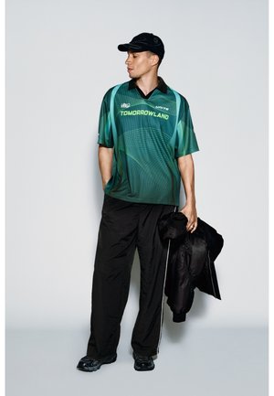 Mann trägt ein grünes Tomorrowland-Shirt, schwarze weite Hose, schwarze Kappe, hält eine schwarze Jacke und steht vor einem einfarbigen hellen Hintergrund.