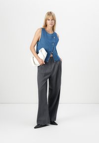 Denim vest met knopen aan de voorkant, gecombineerd met grijze high-waisted wijde broek. Model houdt een witte clutch vast. Eenvoudig ontwerp, strakke lijnen.