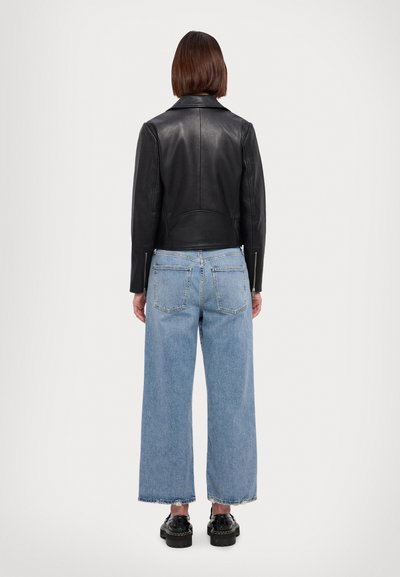 Giacca di pelle nera con un design aderente, abbinata a jeans a gamba larga di colore blu chiaro. La giacca presenta un colletto rotondo e dettagli con zip.