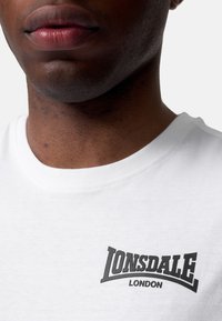 Bijela pamučna majica s okruglim izrezom, koja sadrži crni logo s natpisom "Lonsdale London" na prsima. Glatka tekstura.