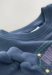 Niebieska bluza z detalami, takimi jak prążkowany okrągły dekolt, mankiety, miękka tekstura oraz graficzny wzór z gwiazdami i kwiatowymi motywami na przodzie.