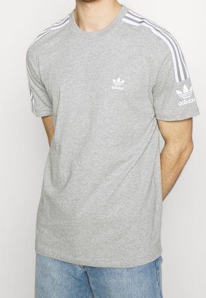 Mężczyzna noszący jasnoszarą koszulkę Adidas z białymi paskami na ramionach oraz logo Adidas na piersi i lewym rękawie.