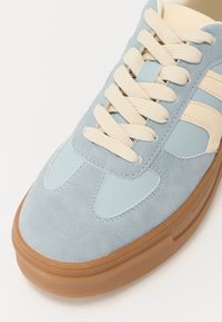 Zapatilla de ante azul pálido con detalles en cuero beige y cordones a juego. Presenta una puntera redonda y suela de goma marrón.