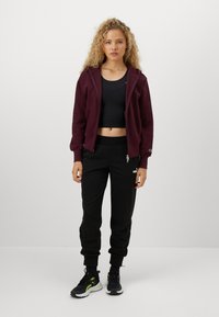 Vinröd zip-up hoodie, svart crop top, svarta joggers med logotyp och svarta sneakers med neondetaljer. Slät textur, avslappnad design.