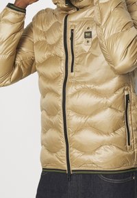 Giacca a piumino color oro metallizzato, con motivo trapuntato, cerniere nere e una tasca frontale. Materiale liscio e testurizzato con polsini elastici.