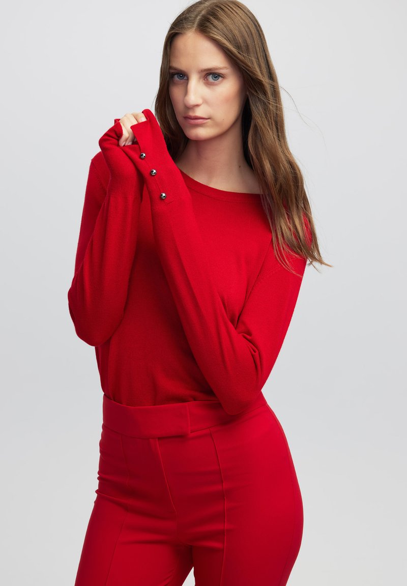 Maglione rosso a maglia con scollatura ampia e maniche lunghe, caratterizzato da dettagli in bottone ai polsini, abbinato a pantaloni rossi su misura.