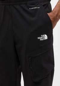 Κοντινή λήψη της μαύρης τσέπης παντελονιού The North Face με φερμουάρ και ετικέτα υφάσματος FLASHDRY-XD, χέρι μερικώς στην τσέπη.