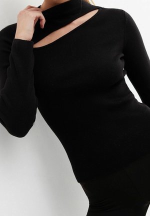 Vrouw draagt een nauwsluitende zwarte trui met lange mouwen en een diagonale uitsnede bij het sleutelbeen, poseert met één hand bij haar nek.