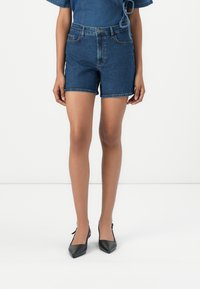 Bermuda de ganga em azul escuro com cintura alta e design de cinco bolsos. Textura suave e barra crua nas aberturas das pernas. Combinada com sapatos de salto pretos.