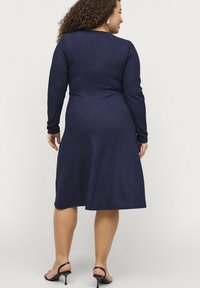 Marineblaues Langarmkleid aus strukturiertem Stoff, eng anliegendem Oberteil, schwingendem Rock und knielanger Länge. Getragen mit schwarzen Absatzsandalen.