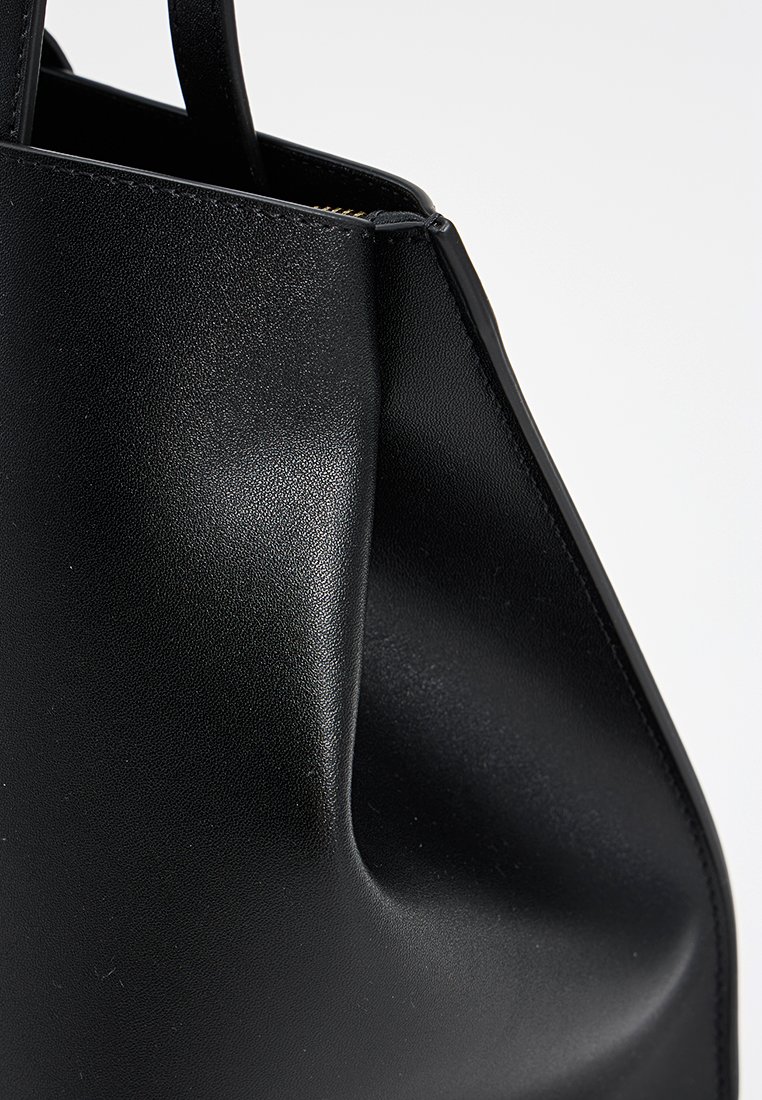 Primo piano di un angolo di una borsa in pelle nera liscia con bordi cuciti e una tracolla su uno sfondo chiaro uniforme.