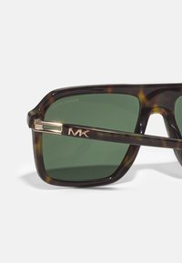 Óculos de sol em padrão tartaruga com lentes verdes, apresentando uma armação retangular elegante e um detalhe em tom dourado na têmpora com gravação "MK".