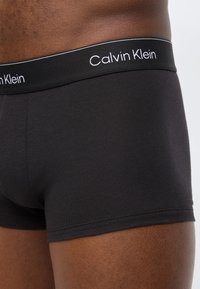 Μαύρα ανδρικά μπόξερ με λογότυπο Calvin Klein στη μέση, που φοριούνται από άτομο με σκούρο δέρμα, εμφανιζόμενα από τη μέση μέχρι το μέσο του μηρού.