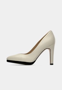 Μπεζ γόβα με ψηλό τακούνι, γυαλιστερή επιφάνεια και μυτερή μύτη. Διαθέτει αντίθετη μαύρη σόλα και τακούνι, δημιουργώντας καθαρό περίγραμμα.