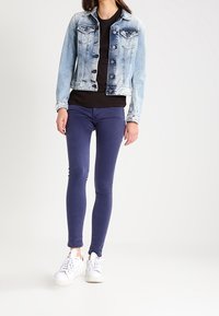 Ljusblå jeansjacka med knäppe och två bröstfickor, lagd över en svart skjorta. I kombination med marinblå smala jeans och vita sneakers.