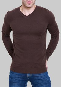 Pull en V marron à manches longues, fabriqué dans un matériau lisse, avec une silhouette ajustée et des poignets et ourlet côtelés. Porté avec un jean bleu.