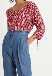 Blusa roja con estampado y mangas abullonadas, con un detalle de botones, combinada con pantalones de mezclilla azul de cintura alta con pliegues y cremallera.