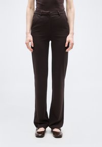 Γυναίκα που φοράει καφέ παντελόνι κανονικής γραμμής με ψηλή μέση και καφέ παπούτσια Mary Jane, σκιαγραφημένη σε απλό λευκό φόντο.