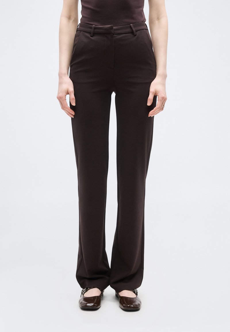 Γυναίκα που φοράει καφέ παντελόνι κανονικής γραμμής με ψηλή μέση και καφέ παπούτσια Mary Jane, σκιαγραφημένη σε απλό λευκό φόντο.