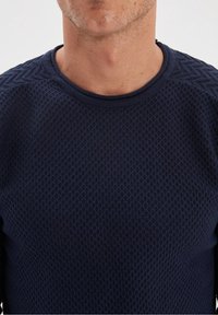 Detailní záběr na mužský krk a horní část hrudníku, oblečeného v tmavě modrém texturovaném kulatém svršku na jednobarevném světlém pozadí.