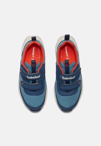 Modré tenisky s textilným a syntetickým zvrškom. S páskami na suchý zips, oranžovými detailmi a bielou gumovou podrážkou. Zobrazené logo Timberland.