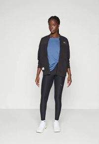 Jachetă sport neagră peste o cămașă albastră și colanți negri, cu o textură fină și accente cu logo-ul Puma. Adidașii albi completează ținuta.