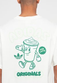 Witte katoenen t-shirt met een groene afbeelding van een lachende ijskoffiebeker, met de tekst "ADIDAS" en "ORIGINALS" in speelse lettertypen.