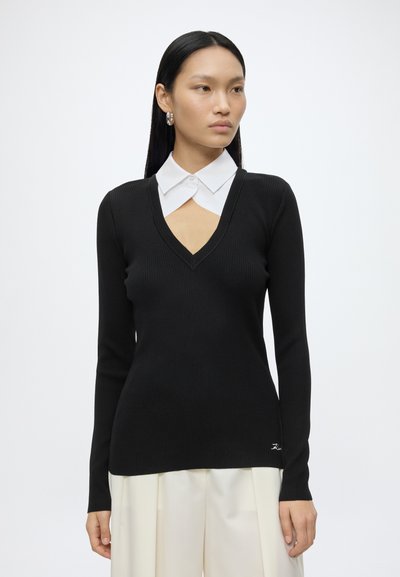 Jeune femme aux longs cheveux noirs porte un pull noir à col en V superposé sur une chemise blanche à col, associé à un pantalon blanc à jambes larges.