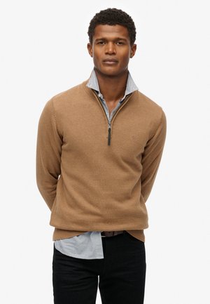 Un pull à col zippé marron en tricot doux, superposé à une chemise à col rayée bleu clair et blanche, associé à un jean foncé.