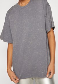 Šedé oversized tričko s texturovaným, mírně žíhaným vzorem. Obsahuje žebrovaný kulatý výstřih a spadlé ramenní švy.