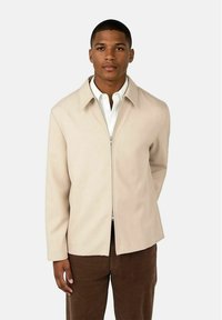 Beige zip-up jacka med spetsig krage och långa ärmar, med en slät textur och minimalisk design, tillsammans med mörkbruna byxor.