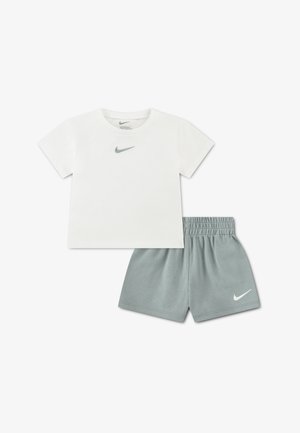 Fehér, rövid ujjú Nike póló szürke pipa logóval, szürke elasztikus derékpántos rövidnadrággal párosítva, amelyen fehér Nike pipa látható.