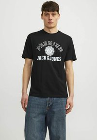 Čierne bavlnené tričko s bielym nápisom "PREMIUM JACK & JONES." Krátke rukávy, okrúhly výstrih a voľný strih. Kombinované s modrými denimovými jeansami.