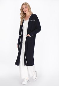 Marineblauer langer Cardigan mit weißen Paspeln, offener Front, zwei Taschen und lockerer Passform. Modell trägt ein weißes Oberteil und weit geschnittene Hosen.