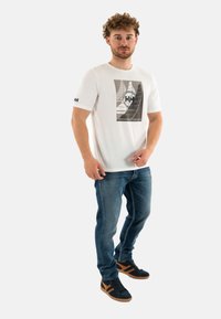 Camiseta blanca de manga corta con un estampado gráfico en tonos de gris y negro, combinada con jeans de mezclilla azul y zapatillas negras.
