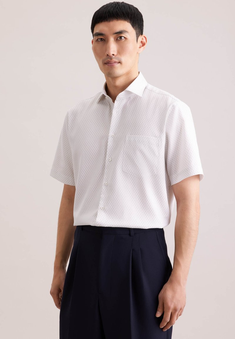 Homme portant une chemise blanche à manches courtes avec des motifs, boutonnée et rentrée dans un pantalon noir taille haute, sur un fond uni.