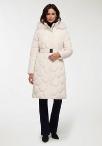 Giacca puffer bianca trapuntata con cappuccio foderato in pelliccia, vita con cintura e chiusure a bottoni a pressione, abbinata a pantaloni scuri e stivali neri.