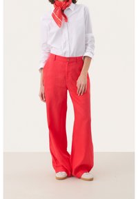 Persoon in een witte button-up shirt, rode wijde broek, rode sjaal om de hals geknoopt, en witte sneakers staand op een effen achtergrond.