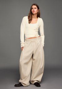 Kremasta majica na preklop s dugim rukavima i detaljem za vezanje, uparena s bež hlačama širokih nogavica. Odjeća je opuštenog kroja i mekanih tekstura.