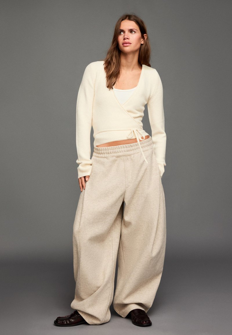 Kremasta majica na preklop s dugim rukavima i detaljem za vezanje, uparena s bež hlačama širokih nogavica. Odjeća je opuštenog kroja i mekanih tekstura.