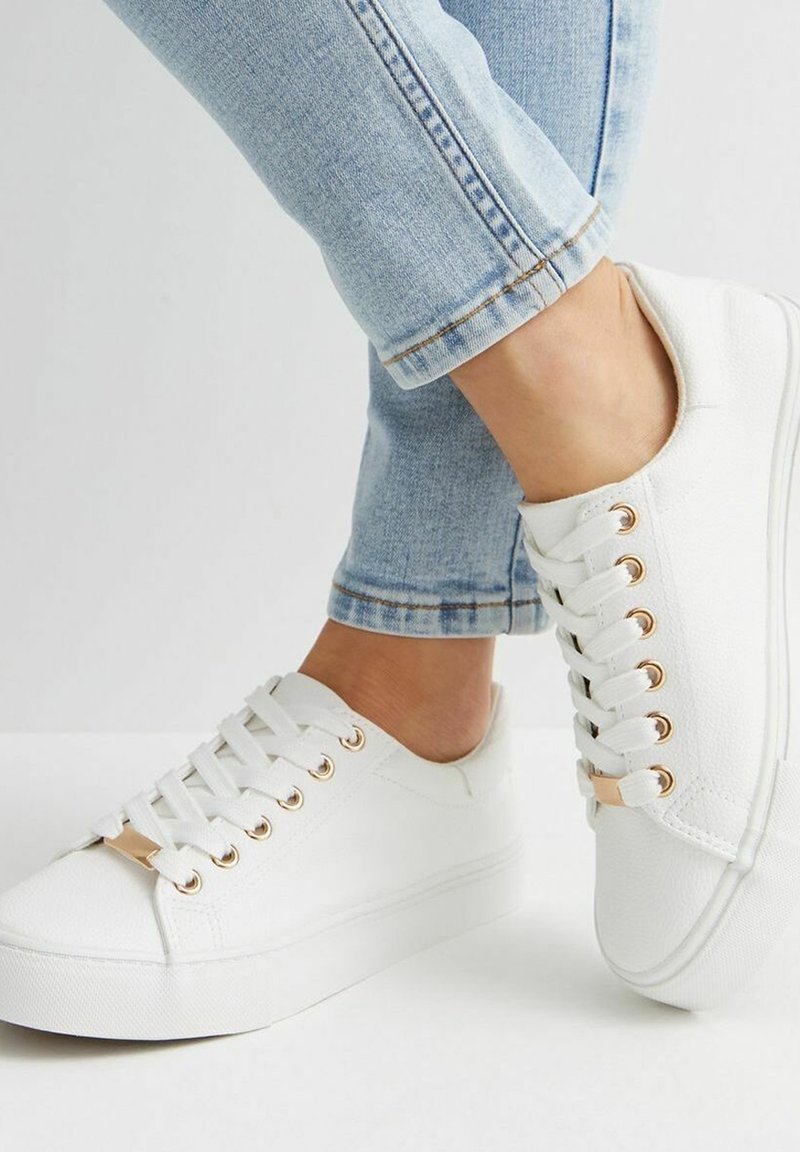 Vita canvas sneakers med guldöglor och en tjock gummisula, parat med ljusblå jeans med uppvikta ben.