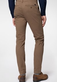 Homme portant un pantalon marron ajusté, des chaussettes grises, des chaussures en cuir marron et une chemise à manches longues bleu foncé, vu de dos.
