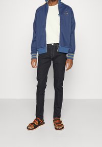 Muž v modré bundě na zip s logem smajlíka, bílém tričku, černých úzkých džínách a černých sandálech s oranžovými pásky, stojící.