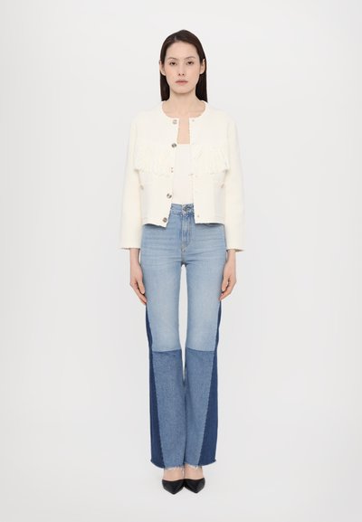 Weißer Fransenjacke mit silbernen Knöpfen, kombiniert mit hellblauen hoch taillierten Jeans, die dunkelblaue Seitenteile und fransige Säume aufweisen.