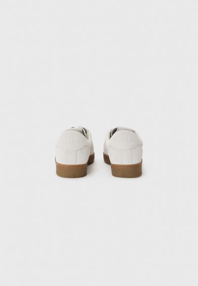 Weiße Wildleder-Sneakers mit einer strukturierten braunen Gummisohle, die abgerundete Fersen und ein minimalistisches Design aufweisen. Keine sichtbaren Logos oder Muster.
