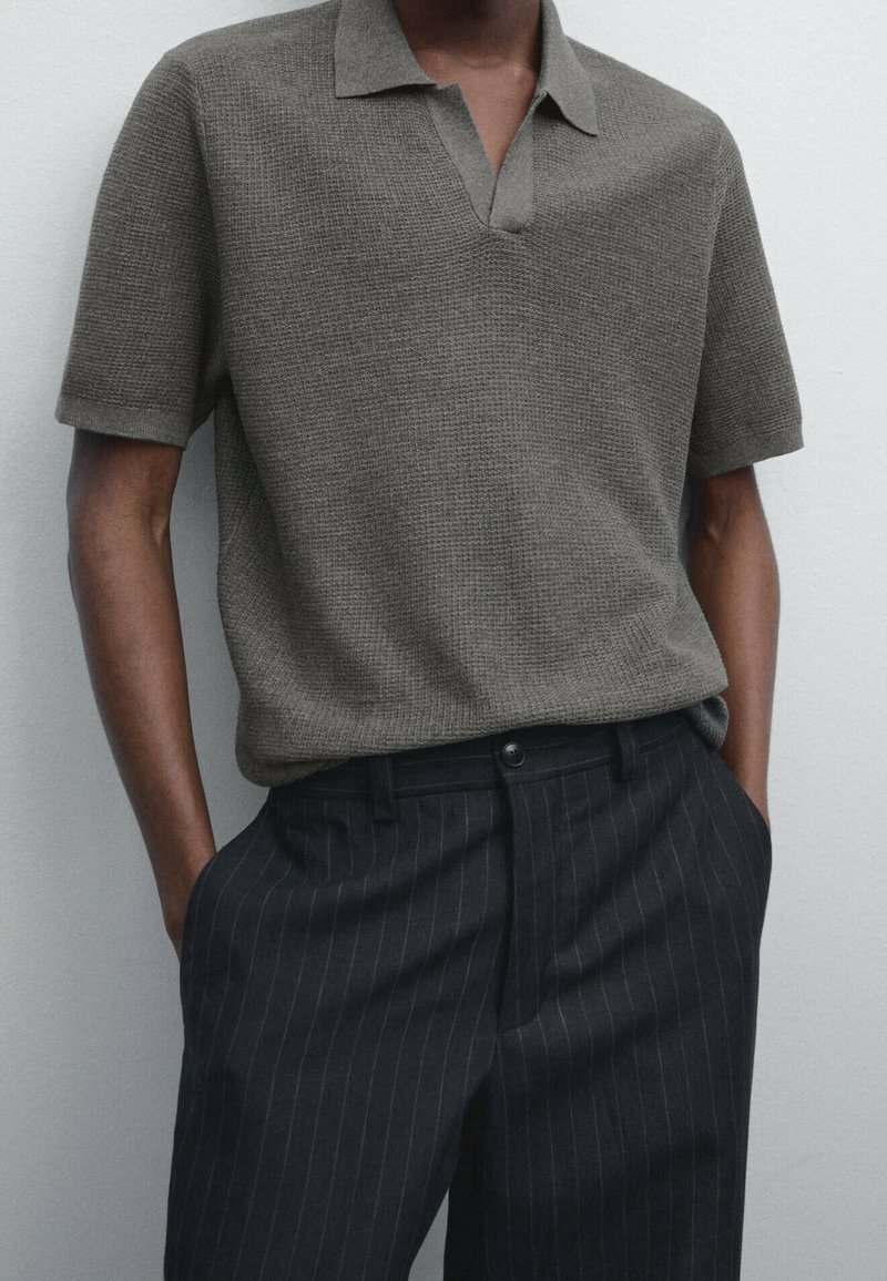Homme portant un polo marron texturé à manches courtes, rentré dans un pantalon sombre à fines rayures, les mains dans les poches.