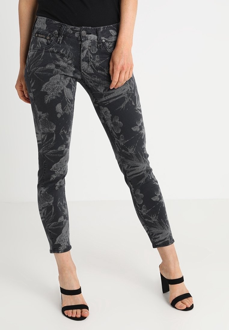 Femme portant un jean skinny noir avec des motifs floraux gris foncé et des sandales à talons noirs sur un fond blanc uni.